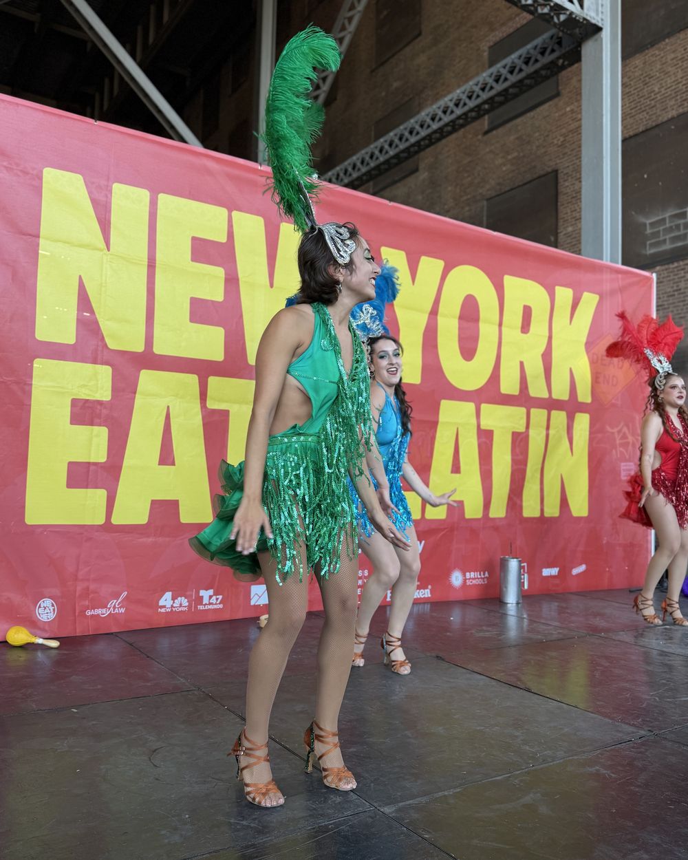 A dancer in green feathers in front of the New York Eats Latin stage banner and the sponsor bar